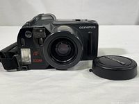 Front view of Olympus Infinity Super Zoom 300 35mm film camera in black with lens extended and Olympus lens cap beside it.