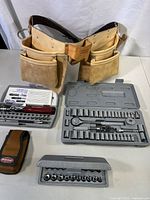 Green cowhide leather tool belt with various storage pockets and metal attachments resting on a white surface along with three sets of driver bits and sockets in plastic cases.