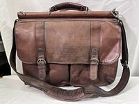 Front view showing brown leather construction, two buckle straps on flap, front pockets, handle and shoulder strap.