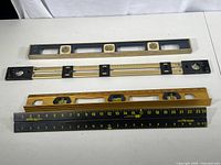 Four measuring and leveling tools laid out on a white surface: one blue-framed spirit level, one yellow Stanley spirit level, a black Stanley measuring ruler, and a metal folding Moore ruler.