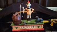 Front view of the cast iron coin bank featuring a clown, dog, hoop and barrel labeled 'Trick Dog'.
