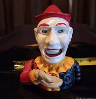 Front view of cast iron clown coin bank showing red hat, red and black outfit, and open hand for receiving coins.
