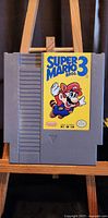 Front view of the original Super Mario Bros 3 Nintendo NES grey cartridge with the game label showing Mario in raccoon suit flying.