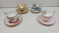Four vintage porcelain teacups and saucers arranged in a row showing different pastel colors and floral, geometric, and plain patterns.
