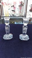 Pair of clear etched crystal candle holders standing on a dark cloth, showing front and side profiles with hexagonal bases and floral etching.