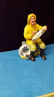 Front view of yellow fisherman's porcelain figurine showing him seated, painting lifebuoy with pipe in mouth