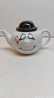 Front view of the white ceramic teapot with smiley face and black dome lid on top.