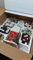 View of multiple porcelain Christmas village buildings in original styrofoam packaging inside cardboard box showing multicolor holiday-themed designs including snow, trees, and wreaths.