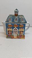 Front view of multicolour ceramic teapot shaped as a house with blue roof and floral window boxes