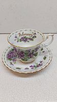 Teacup and saucer set showing floral violet pattern with February inscription inside the cup and gold rims.