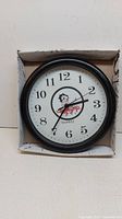 Front view of the round black plastic wall clock showing full clock face with Betty Boop image, numbers and hands inside a worn cardboard box.