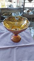 Side view of vintage iridescent amber carnival glass compote showing the pedestal base and flared scalloped rim with grape and leaf embossed pattern.