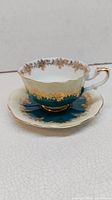 Teal and gold patterned porcelain teacup and saucer with gold floral accents, scalloped edges, and gold-trimmed handle.