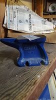 Blue painted metal anvil showing full length with a square horn and flat top surface, positioned on wooden surface with newspaper behind.
