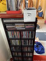 Wooden CD rack filled with jewel-case CDs across seven shelves and one crate of loose CDs on top