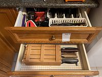 Two kitchen drawers showing organized cutlery and utensils including a wooden knife block with knives placed horizontally, and a tray with forks, spoons, knives, and assorted small kitchen tools.