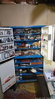 Metal cabinet with blue shelves containing various small parts and supplies, doors open to show inside and compartments on door