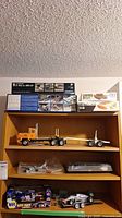 Shelf with multiple boxed model kits including an F-14A vs MiG-29 aircraft kit, a small white and green box, and assembled orange construction truck with trailer.
