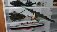 Shelf showing military helicopter models, tank, plane, and RMS Queen Mary ship model together