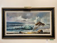 Full view of the framed ocean wave painting showing waves crashing on rocks with cloudy sky and distant mountains.