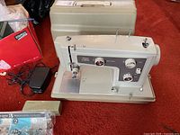 Full view of Sears Kenmore portable sewing machine, beige color, with foot pedal and beige carrying case, set on red carpet.