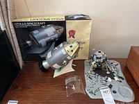 Photo showing the Apollo Spacecraft Command Service Module with display box behind and moon lander diorama on wooden surface, with clear plastic canopy piece in front.