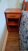 Front view of the Vilas maple nightstand showing the drawer and two open shelves, next to a bed with a patterned comforter.