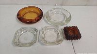 Top view showing five assorted glass ashtrays including round amber, round clear, two clear square, and one textured brown square ashtray.