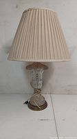 Full front view of a glass table lamp with beige pleated shade on a white surface against a white wall.