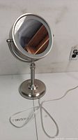 Front view of round lighted makeup mirror with metal stand on white surface, cord visible