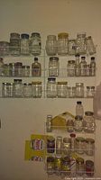 Photo showing multiple wire racks mounted on wall holding assorted clear glass mason jars and bottles with various lid colors including silver, gold, blue, white, and black. The jars vary in sizes and some have textured designs on the glass. There are also a couple of paper packets stored on lower rack.