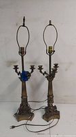 Pair of brass candelabra-style table lamps showing full height and base. One lamp has a blue tape holding a piece in place and the harp frame is bent on one lamp.