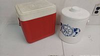 Photo showing both the vintage cooler and the ice bucket side by side on a white surface, cooler is red and white plastic, ice bucket is white with blue design.