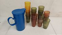 Blue plastic pitcher with handle and lid, accompanied by ten assorted colored vintage plastic glasses.