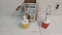 Two vintage Ray-O-Vac portable table lamps shown side by side; one yellow-based lamp with original box behind it, and another orange-based lamp with a white lampshade to the side.