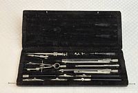 Photo of antique drafting instruments arranged neatly in original leather case with lid open, showing metal drafting tools including compasses, dividers, ruling pens, and other accessories secured inside.