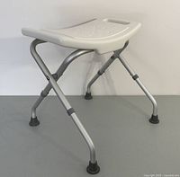 Side view of folding bath chair showing seat surface and tubular metal legs with rubber feet.