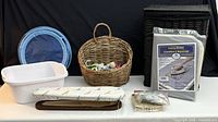 Black wicker hamper with cloth liner, round blue folding hamper, light brown wicker basket with clothespins, new ironing blanket in package, sleeve ironing board, assorted clothespins and small baskets all shown on white table against black background.