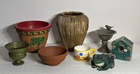 Overview of all garden decor items: taupe bird bath, green distressed bird house, plastic frog, wood planter cover, painted clay pot, green urn planter cover, terracotta pot, and decorative cup.