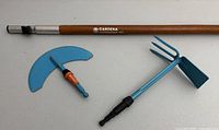 Wooden handle with two blue metal attachments (hand hoe with three prongs and flat blade, and curved edger) laid out side by side showing components separately.