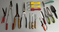 Wide shot showing all gardening hand tools laid out on a gray surface, including pruning shears, weeding tools, trowels and rake, showing their sizes, handle colors, and condition.
