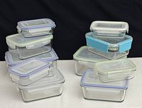 Stacked arrangement of 11 Glass Lock rectangular and square food storage containers of various sizes with plastic lids in blue, green, and purple clips on a white surface against dark background.