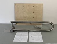 Fold down toilet assist rail mounted on plywood wall plate with installation hardware and manuals on a table surface.