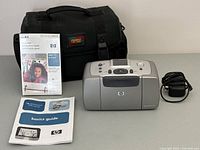 Photo showing the HP Photosmart 245 photo printer, Optex carry case, power adapter, HP photo paper, and basics guide manual.