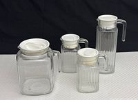 Four clear ribbed glass jars with glass handles and white lids arranged on a white surface against dark background, showing variety in sizes and ribbed texture.