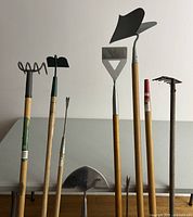 Six long-handled garden tools standing upright against a table: spiral coil cultivator, small hoe, dandelion spike, triangular hoe, rectangular edger, ridge plow