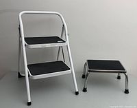 Foldable two-step metal step stool next to low single-step metal stool on floor.