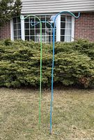 Blue and green metal hanging basket brackets standing in front of bushes and a brick house window.