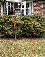Pair of tall red metal stakes with double hooks for hanging baskets, standing on grass with green bushes and a house window in the background.