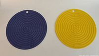 Pair of round rubber trivets, one blue and one yellow, showing the spiral maze pattern and hanging hole.